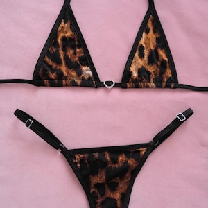 Op de afbeelding: Een tweedelige bikiniset met een luipaardprint in bruin en zwart. De top is een driehoekmodel met een kleine hartvormige versiering. De onderkant is een stringmodel met verstelbare bandjes. De set wordt gepresenteerd op een roze achtergrond.