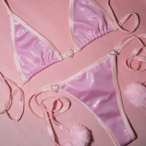 Könnte beinhalten: Ein hellrosa Bikini-Set mit einem strukturierten, schimmernden Stoff. Das Triangel-Top und der Tanga-Slip sind mit Weiß eingefasst und mit herzförmigen Verzierungen versehen. Rosa Pompons sind an den Bindebändern befestigt, alles vor einem rosa Hintergrund.