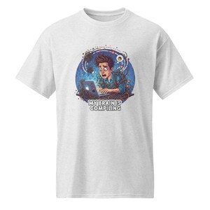 May include: A light gray t-shirt featuring a graphic of a person with a shocked expression working on a laptop, surrounded by tangled wires and blue circuit patterns. The text "MY BRAIN IS COMPILING" is printed in bold white letters below the illustration.
