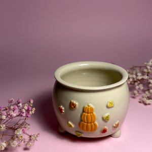 Puede incluir: Una maceta de cerámica color crema con forma redondeada y tres pequeños pies. La maceta está decorada con una calabaza naranja en relieve y coloridas hojas de otoño. El fondo es de color rosa liso.