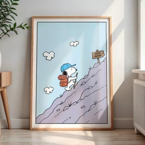 May include: Framed print of Snoopy, wearing a blue cap and carrying a backpack, climbing a mountain. A sign at the summit reads "KEEP GOING". The background is a light blue sky with white clouds. The artwork is in a wooden frame.