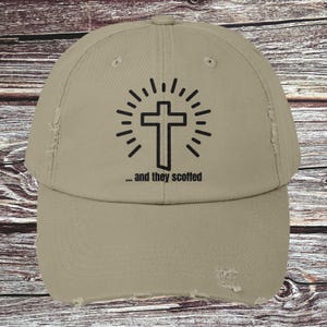 May include: A tan baseball cap with a distressed finish. The cap features a black cross design with radiating lines and the text "... and they scoffed" in black.