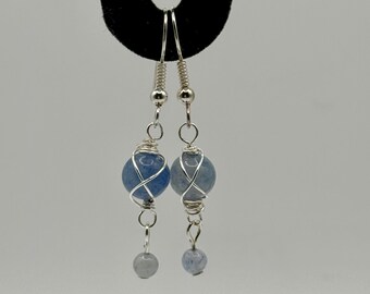 May include: Dangle earrings with blue and silver beads. Each earring has a large, round, blue bead wrapped in silver wire, with a smaller, grey bead dangling below. The earrings hang from silver-coloured hooks.