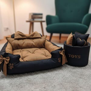 May include: A dog bed with a black and tan color scheme, featuring a bow and paw print details. A matching toy basket with the word "TOYS" is next to the bed. The bed has a soft, cushioned interior.