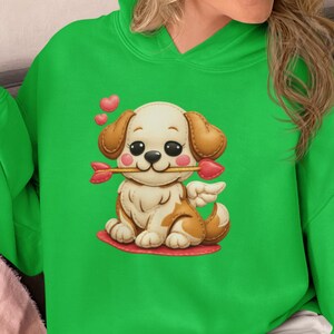 以下が含まれることがあります： 明るい緑色のパーカー。漫画の子犬のデザインで、ハートの先端が付いた矢を口にくわえ、小さな翼があります。子犬は赤いハートの上に座っています。パーカーは鮮やかな緑色です。