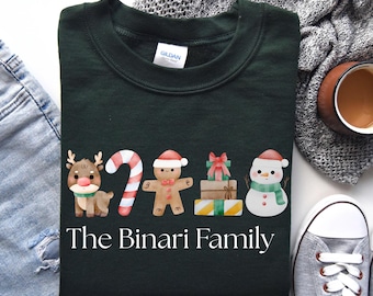 May include: Dark green sweatshirt with a festive design featuring a reindeer, candy cane, gingerbread man, wrapped gifts, and a snowman. The text "The Binari Family" is printed below the illustrations. The sweatshirt is made of a soft material.