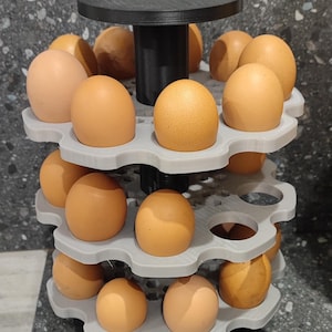 Puede incluir: Un soporte para huevos de tres niveles hecho de plástico gris, con una docena de huevos marrones. El soporte tiene una base y una parte superior negras y está diseñado para mantener los huevos organizados. Los huevos son de color marrón claro.