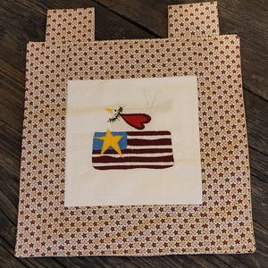 May include: A patriotic, rectangular fabric item with a cream-colored center featuring an embroidered American flag and a stylized bird. The border and tabs are patterned with red stars on a cream background.