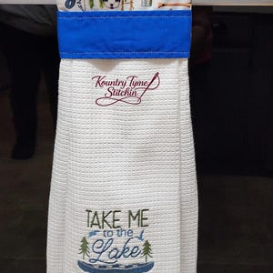 May include: White kitchen towel with a blue border and fishing-themed fabric. The towel has the text "Take Me to the Lake" and "Kountry Tyme Stitchin" embroidered on it. A wooden sign that says "Country Kitchen" is above the towel.