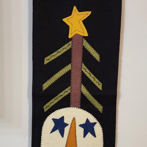 May include: A black felt Christmas wall hanging with a snowman design. The snowman has blue star eyes, an orange carrot nose, and blue button mouth. A yellow star tops the tree. The banner is suspended from a wooden rod.
