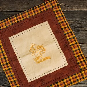 May include: A square Halloween-themed pot holder with a plaid border in yellow, orange, and brown. The center features a cream-colored square with an embroidered design of two skulls and the words "Happy Halloween."