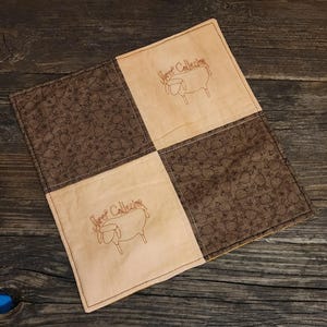 May include: Square, quilted fabric coaster with a patchwork design. Two squares feature a brown sheep pattern, and two squares are beige with a sheep illustration and the text "Sheep Collection" embroidered in brown. The coaster is on a wooden surface.