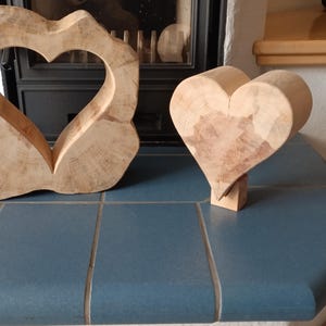Könnte beinhalten: Zwei Holzskulpturen in Herzform. Eine ist ein massives Herz auf einem kleinen Sockel. Die andere ist ein herzförmiger Ausschnitt in einer größeren, abstrakten Form. Beide sind aus hellem Holz gefertigt und stehen auf einer blauen Fliesenoberfläche.