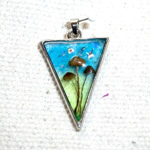 Puede incluir: Colgante triangular con marco plateado. En el interior, un paisaje en miniatura presenta setas marrones sobre un fondo azul y verde, con pequeños detalles en forma de estrella. El colgante está diseñado para ser usado como joya.