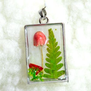 Puede incluir: Colgante rectangular con marco plateado. En el interior, una seta roja y un helecho verde están encapsulados en resina transparente. El colgante tiene un lazo plateado para una cadena.