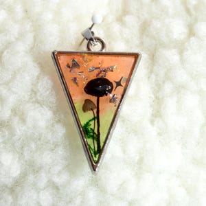 Puede incluir: Colgante triangular con marco plateado. En el interior, una escena en miniatura presenta una seta negra, setas marrones más pequeñas y follaje verde sobre un fondo naranja y amarillo. El colgante incluye una argolla plateada y un alfiler blanco.