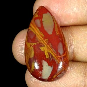 Könnte beinhalten: Ein polierter, tropfenförmiger Edelstein. Der Stein zeigt eine lebendige Mischung aus Farben, mit einer Basis aus tiefem Rot, akzentuiert durch Adern und Flecken in Gelb, Beige und Creme. Die Oberfläche ist glatt und reflektierend.