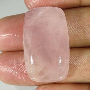 Puede incluir: Una piedra preciosa de cuarzo rosa pulida, de forma rectangular. La piedra tiene un tono rosa suave y translúcido con sutiles vetas internas. La forma es un rectángulo redondeado, ideal para la joyería o la colección.