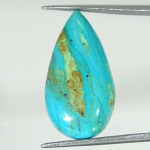 May include: A teardrop-shaped turquoise gemstone with a polished surface. The stone features a vibrant blue hue with streaks of tan and brown. Fine black lines and speckles add detail to the natural stone.