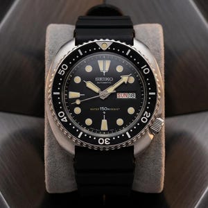 May include: A Seiko automatic wristwatch with a black dial, luminous hour markers, and a black rotating bezel. The watch features a day-date window at the three o'clock position, silver-tone hands, and a black rubber strap. The dial displays the text Seiko, Automatic, and Water 150m Resist.
