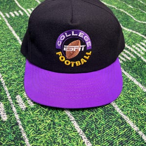 May include: Black and purple baseball cap with the text "COLLEGE FOOTBALL" and the ESPN logo. The cap has a purple brim and button, and the text is in purple and gold. The hat is likely a sports fan accessory.