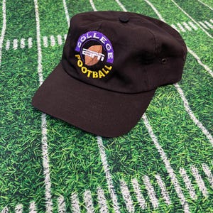 May include: A black baseball cap with a curved brim. The cap features a circular logo with the text "COLLEGE FOOTBALL" in yellow and the ESPN logo in the center. The background is a green football field.