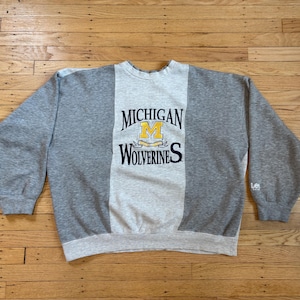 May include: A gray and light gray sweatshirt with the text "MICHIGAN WOLVERINES" in black and yellow. The sweatshirt has long sleeves and a crew neck. The Lee logo is on the sleeve.