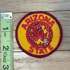 May include: A circular patch with the words "ARIZONA STATE" in maroon lettering. The patch features a yellow center with a maroon border. A red devil with a trident is in the center. The patch is approximately 2 inches in diameter.