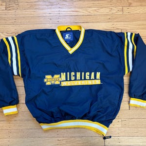 May include: A navy blue Starter pullover windbreaker jacket featuring yellow and white striped trim on the collar, cuffs, and hem. The front displays the yellow text Michigan Wolverines with a block M logo and a Starter brand tag at the neckline.