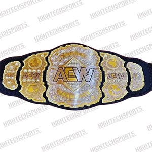Könnte beinhalten: Ein gold-silberner AEW World Championship-Gürtel mit einem schwarzen Lederriemen. Der Gürtel zeigt das AEW-Logo, die Worte "WORLD CHAMPION" und dekorative Goldplatten mit verschiedenen Designs. Der Hintergrund ist weiß.