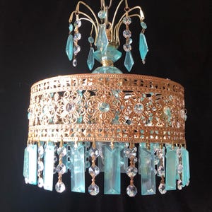 May include: A vintage chandelier with a gold-colored filigree shade and hanging blue-green crystal accents. The chandelier has a decorative top with additional crystal embellishments, creating a glamorous lighting fixture.