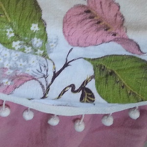 May include: A white fabric tablecloth with a floral print of pink and green leaves and white flowers. The tablecloth has a white trim with white pom-poms along the bottom edge.