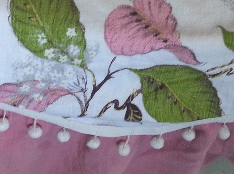 May include: A white fabric tablecloth with a floral print of pink and green leaves and white flowers. The tablecloth has a white trim with white pom-poms along the bottom edge.