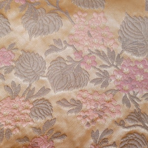 May include: A close-up of a beige fabric with a repeating pattern of pink and gray flowers. The fabric has a textured, woven look.