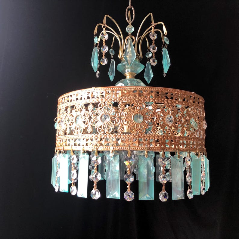 May include: A vintage-style chandelier with a gold-colored metal frame and turquoise-colored glass accents. The chandelier features a decorative filigree design around the shade and hanging crystal-like embellishments. The fixture is suspended from a chain.