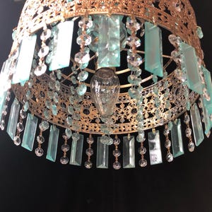 May include: Ornate gold-toned chandelier with hanging rectangular and round crystal accents. The chandelier features a detailed filigree design and a central light fixture. The rectangular crystals are a light teal color.