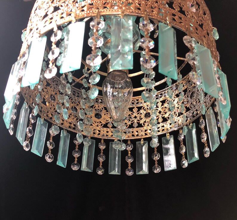 May include: Ornate gold-toned chandelier with hanging rectangular and round crystal accents. The chandelier features a detailed filigree design and a central light fixture. The rectangular crystals are a light teal color.