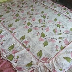 May include: A white bedspread with a pink and green floral pattern. The bedspread has a pink ruffle trim and a white pom-pom trim.