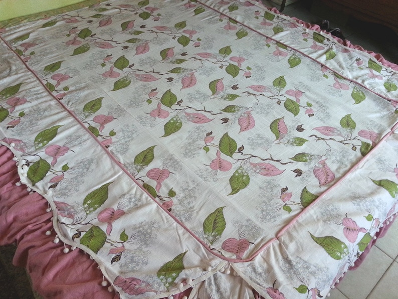 May include: A white bedspread with a pink and green floral pattern. The bedspread has a pink ruffle trim and a white pom-pom trim.