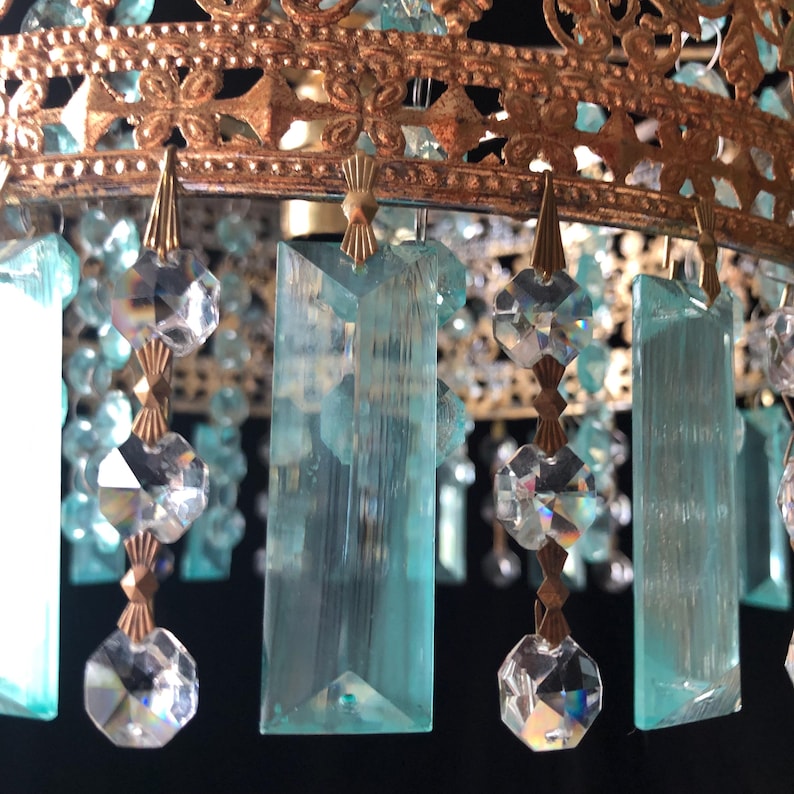 May include: Close-up of a vintage chandelier with ornate gold-colored metalwork. The chandelier features hanging clear and light blue crystal accents. The crystals are various shapes, including rectangular and faceted, adding sparkle and elegance to the design.