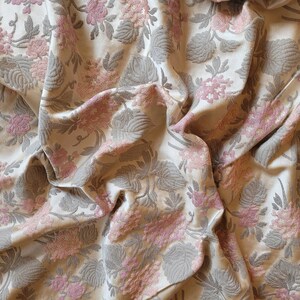 May include: A close-up of a beige fabric with a pink and gray floral pattern. The fabric is wrinkled and has a textured surface.
