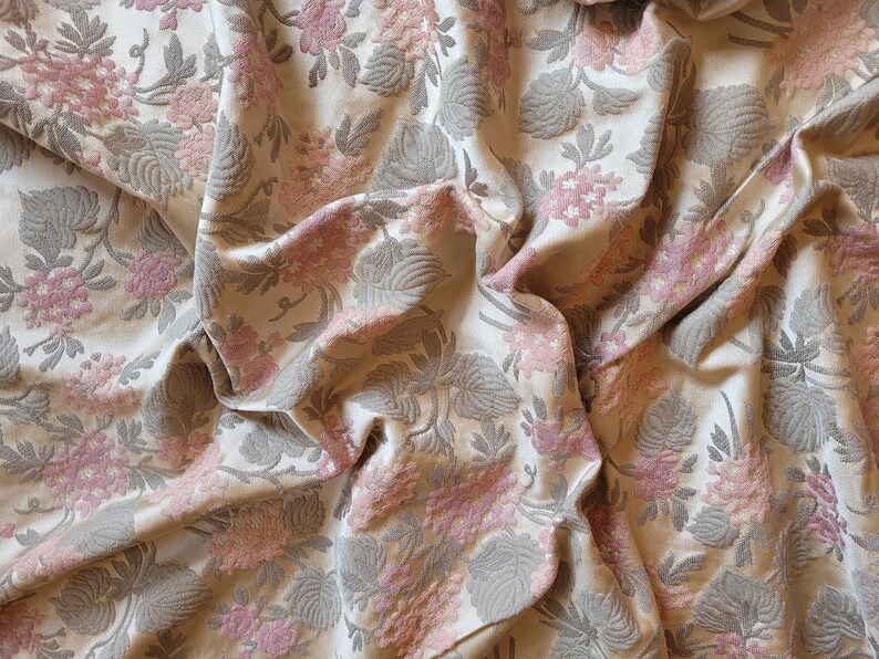 May include: A close-up of a beige fabric with a pink and gray floral pattern. The fabric is wrinkled and has a textured surface.
