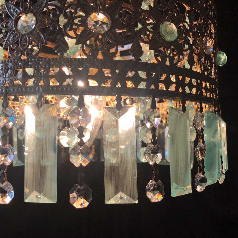 May include: Close-up of a decorative chandelier with a dark metal filigree shade. The chandelier features clear and light green crystal accents and hanging rectangular and faceted crystal pendants. The light shines through the crystals.