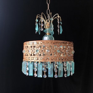 May include: A vintage-style chandelier with a gold-colored metal frame and turquoise-colored crystal accents. The chandelier features a decorative filigree design around the shade and hanging crystal pendants. The fixture is suspended from a chain against a black background.