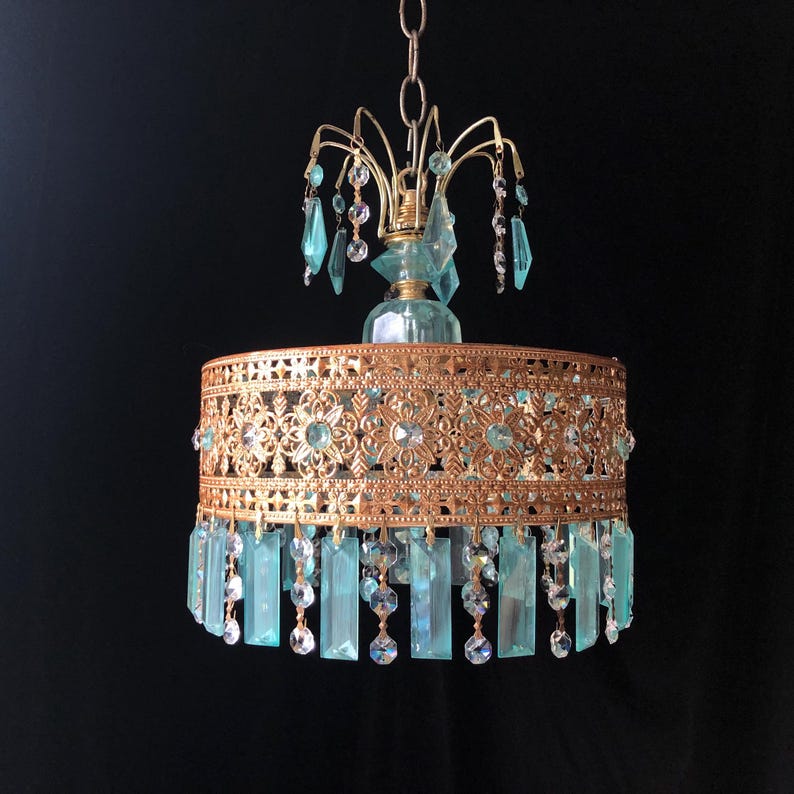 May include: A vintage-style chandelier with a gold-colored metal frame and turquoise-colored crystal accents. The chandelier features a decorative filigree design around the shade and hanging crystal pendants. The fixture is suspended from a chain against a black background.