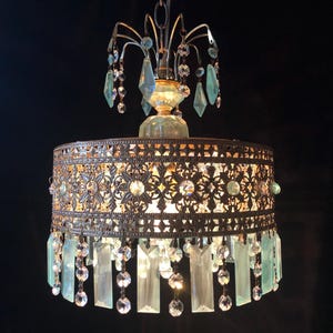 May include: An ornate chandelier with a bronze-colored metal frame and hanging crystal accents. The fixture features a decorative filigree shade with clear and light blue crystal pendants and droplets. The chandelier is suspended from a chain against a dark background.