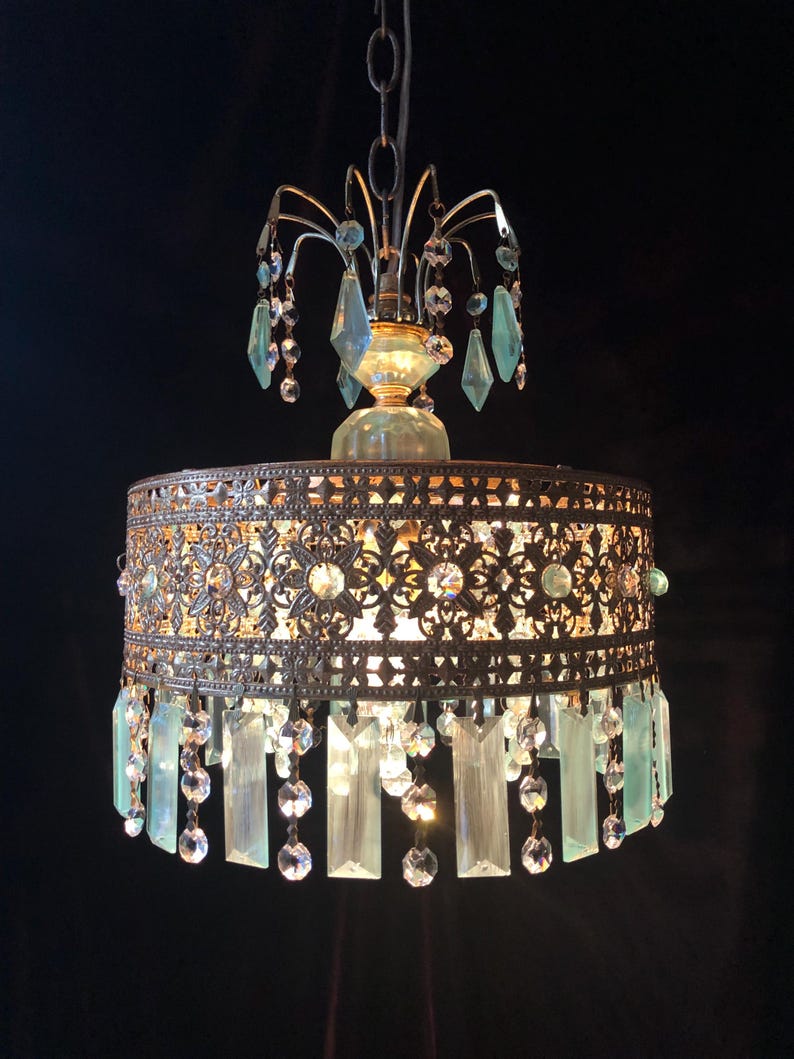 May include: An ornate chandelier with a bronze-colored metal frame and hanging crystal accents. The fixture features a decorative filigree shade with clear and light blue crystal pendants and droplets. The chandelier is suspended from a chain against a dark background.
