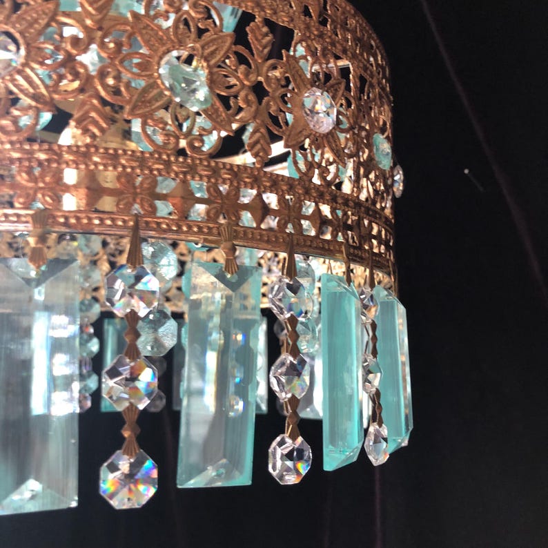 May include: Ornate, antique-style chandelier with a bronze-colored metal frame. The frame features intricate floral and geometric cutouts. Hanging from the frame are clear and light blue crystal pendants and teardrops, creating a sparkling effect. The chandelier is a decorative lighting fixture.