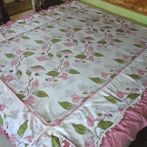 May include: A white bedspread with a pink floral pattern and a pink ruffled border. The bedspread has a white lace trim and white pom-poms along the border.