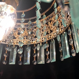 May include: Close-up of a gold-toned chandelier with intricate filigree detailing. The chandelier features hanging rectangular and round aquamarine-colored crystals. A bright light bulb is visible at the top, illuminating the crystals and gold frame.